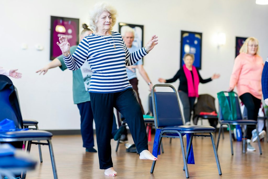 Een oudere dame doet enthousiast mee met de seniorengym, met stoelen in de zaal